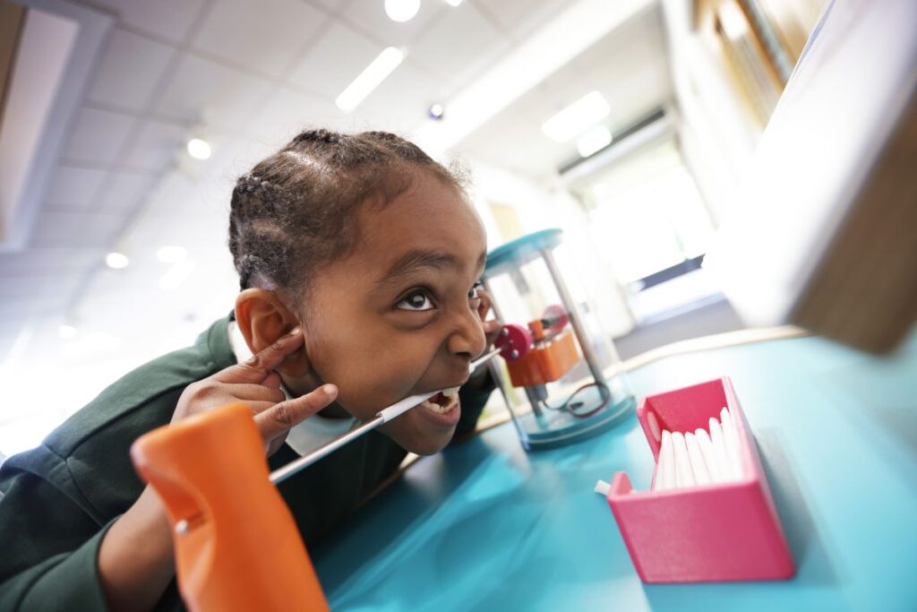 A school-going child with teeth over metal rod covered in a paper straw at science exhibit demonstrating how one can hear through one's teeth