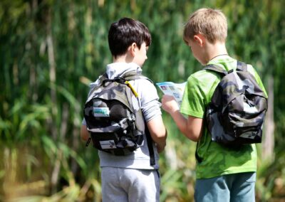 Two boys dressed in T shirts on a sunny day, with backpacks on their backs, are looking at a brochure. The background is green outdoors
