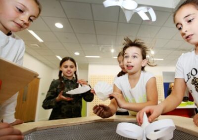 Two children lean over a table blowing air up from below, paper cones and hoops are flying up