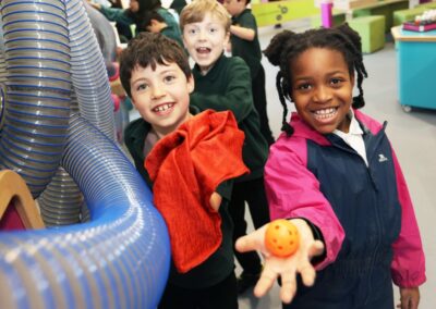Three primary school children, smiling, pose for camera holding up a cloth and ball in front of a wall with tubes mounted.