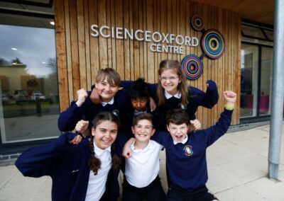 Six pupils pose in front of building with Science Oxford Centre logo on wall. They are smiling, have arms interlinked and are raising free arms in cheer.