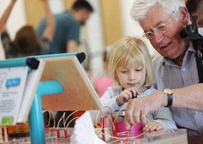 An elderly man, seated, and young girl standing play with wires part of a science exhibit