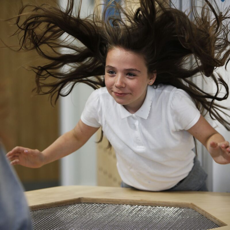 A girl, smiling, leans over a table blowing air up from below. Her long hair blows in the wind.