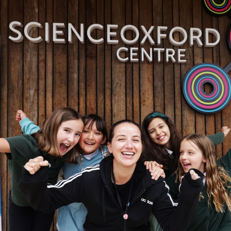 A group of children and a teacher pose for camera and lift their arms and cheer in front of a building with Science Oxford Centre logo behind.