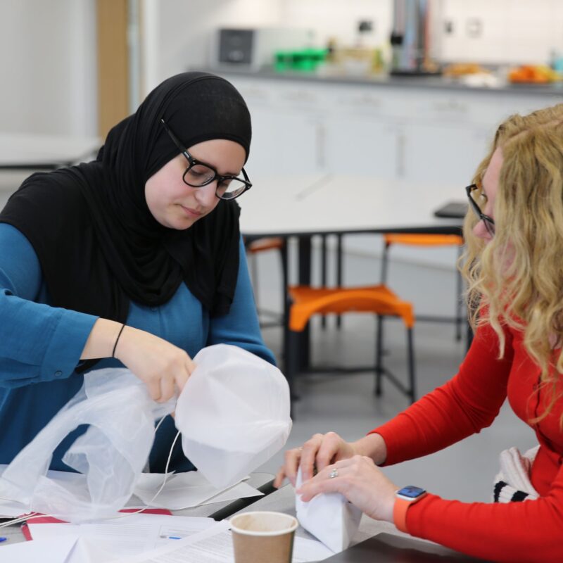 A woman seated at a table uses a plastic bag and string to build something.