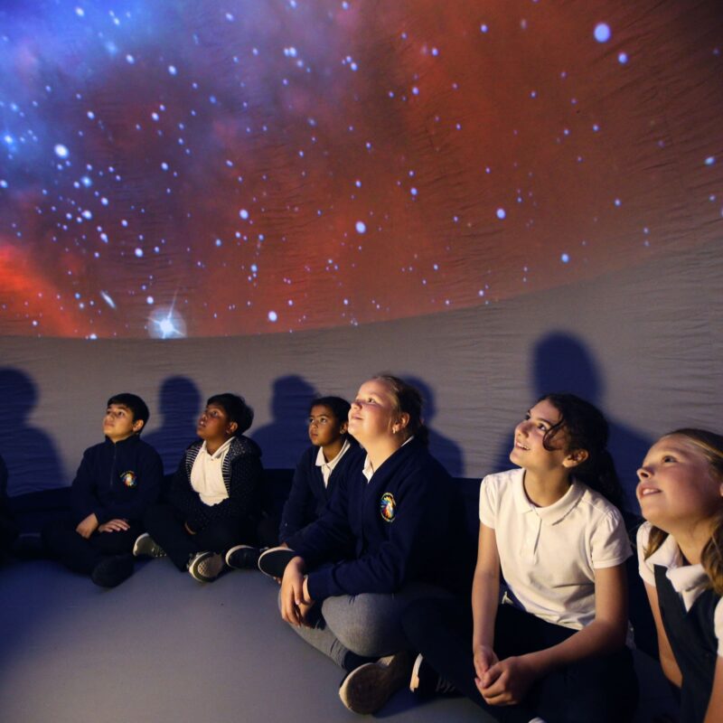 A group of primary school children look at the illuminated celling as part of a planetarium show.