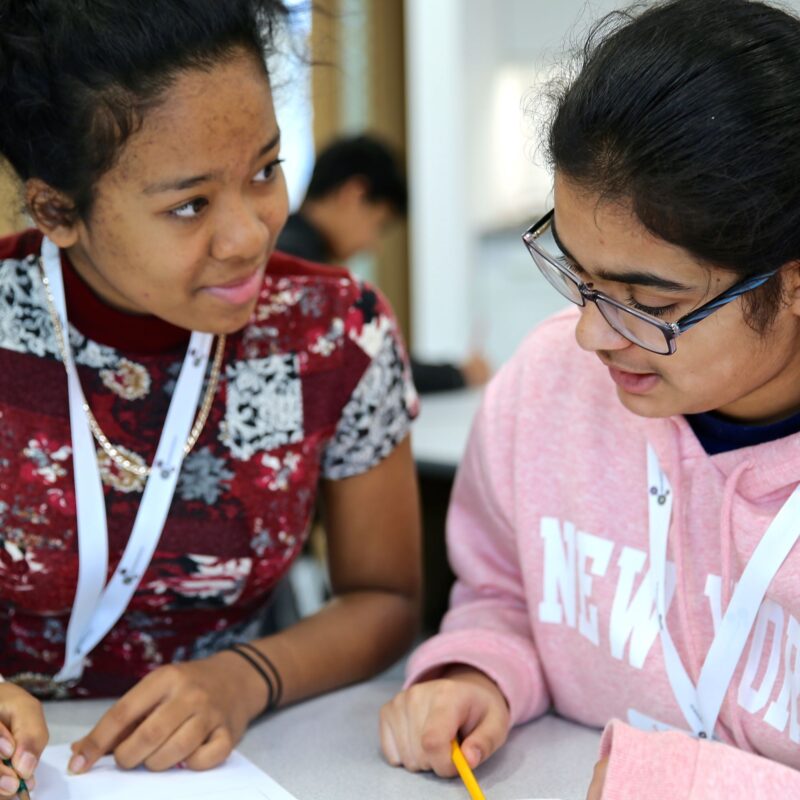 Two secondary school-aged girls are seated at a table and talking, writing something down on paper