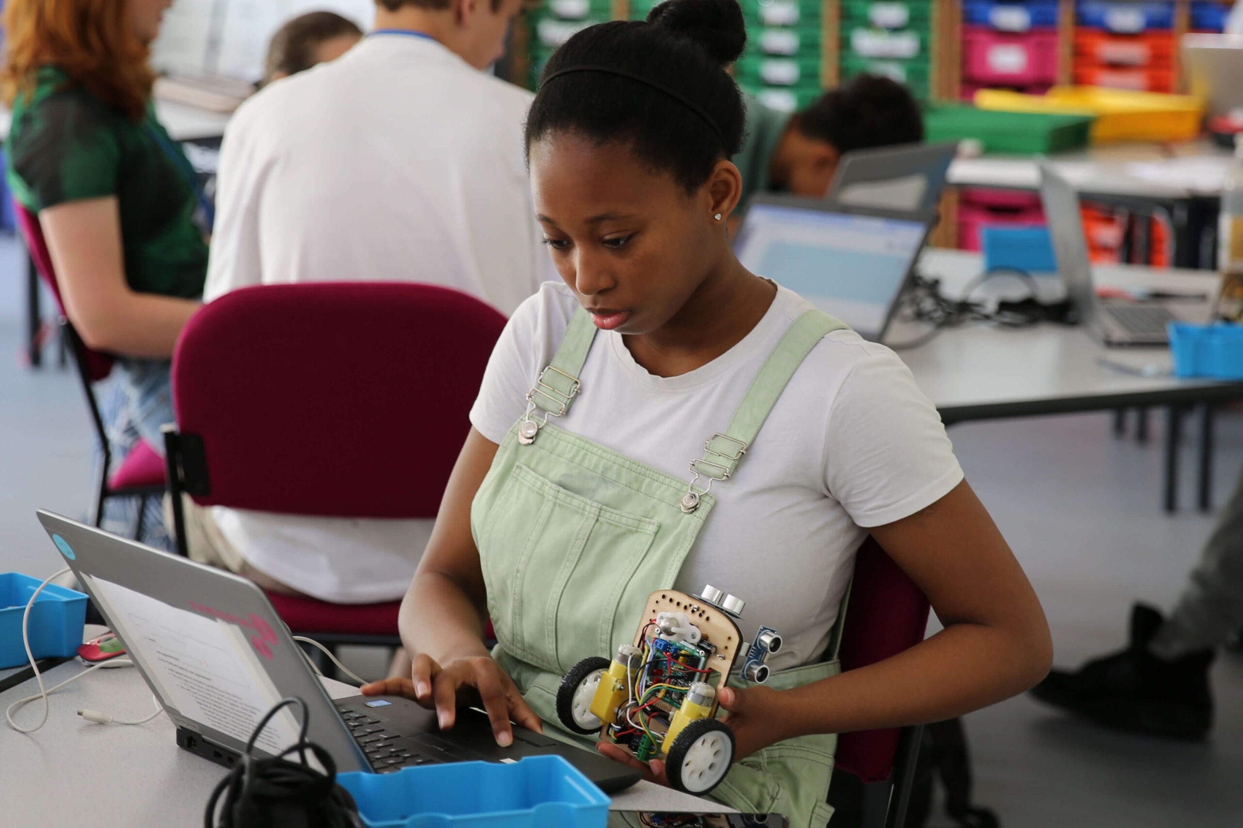 A girl programs a robot with wheels using a laptop which she is looking at.