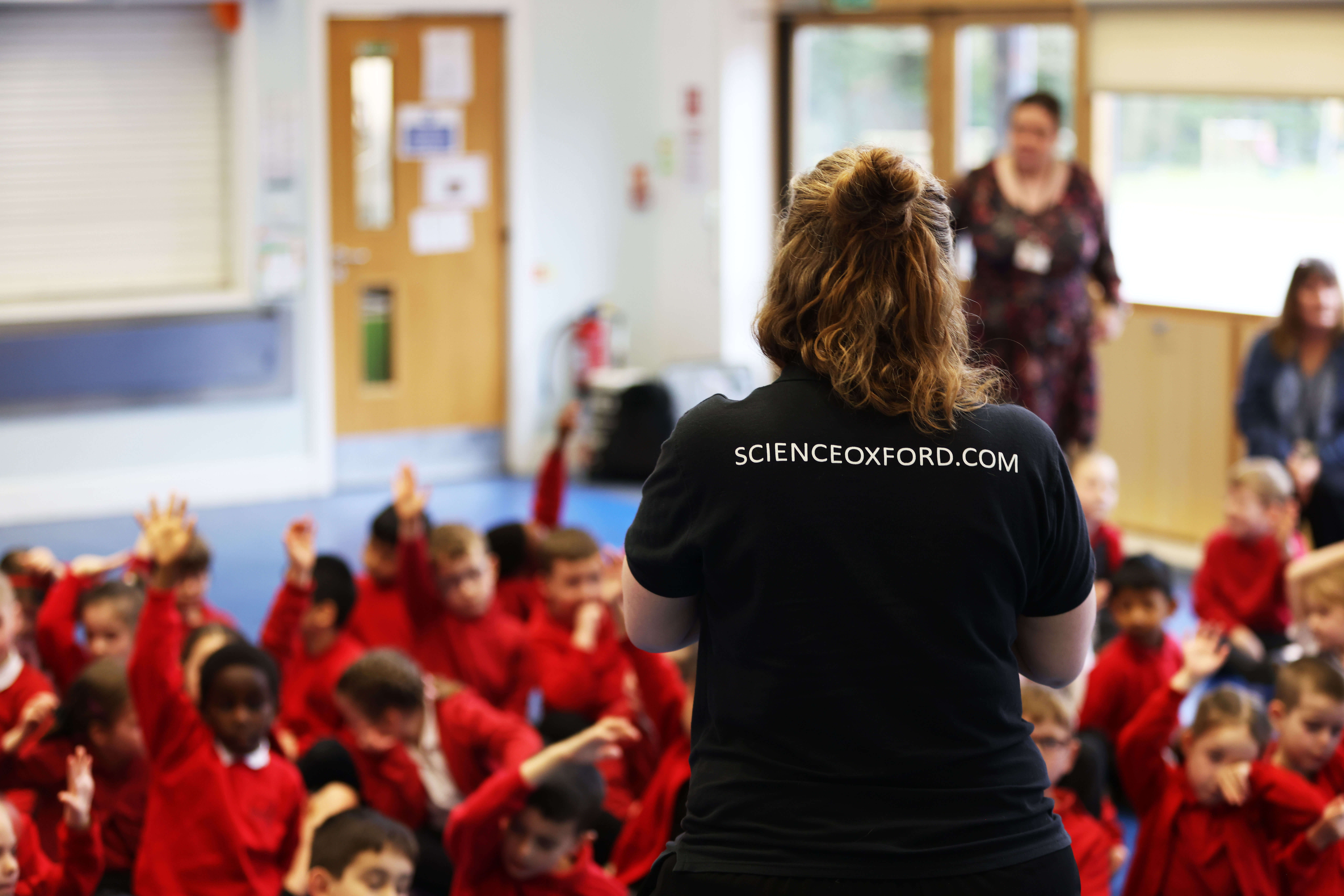 A woman stands with her back to the camera, and her black t-shirt has 'scienceoxford.com' on the back. She is addressing a group of primary school children.