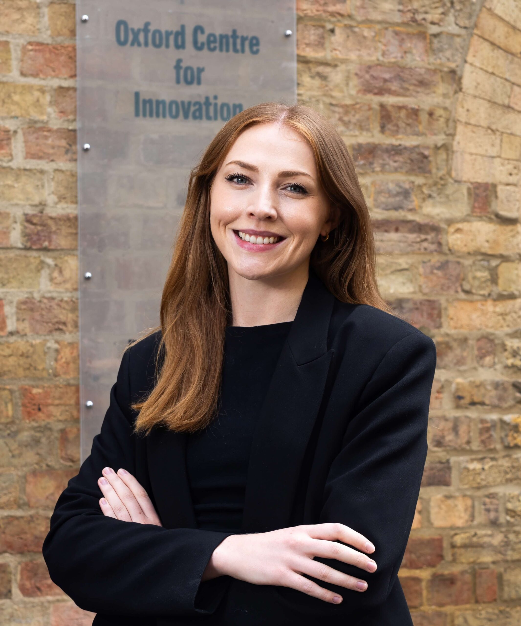 A woman in a black jacket, smiles looking at the camera. She stands outside at building with a sign that reads 'The Oxford Centre for Innovation'.