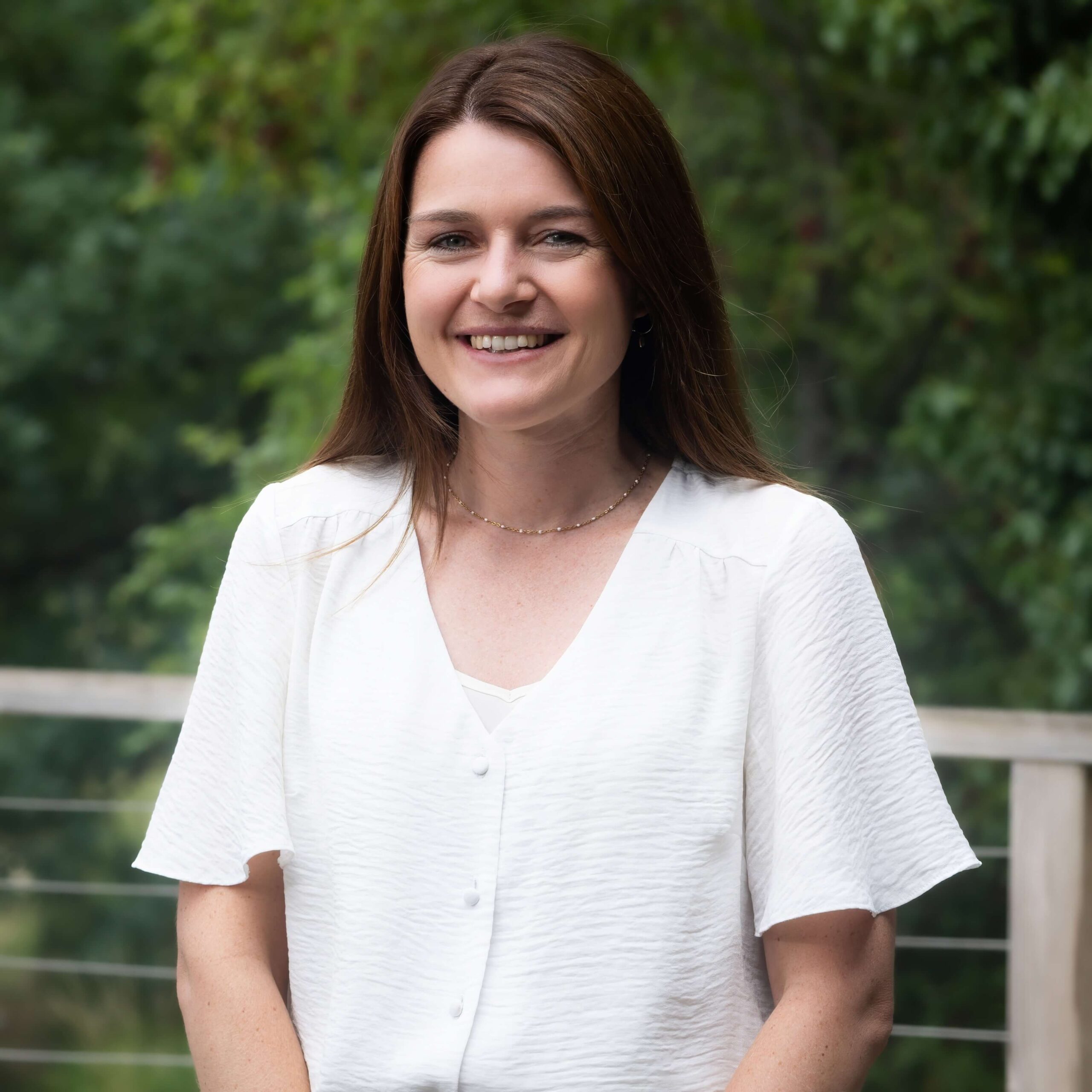 A portrait of a woman in a white blouse standing outside with trees in the background.