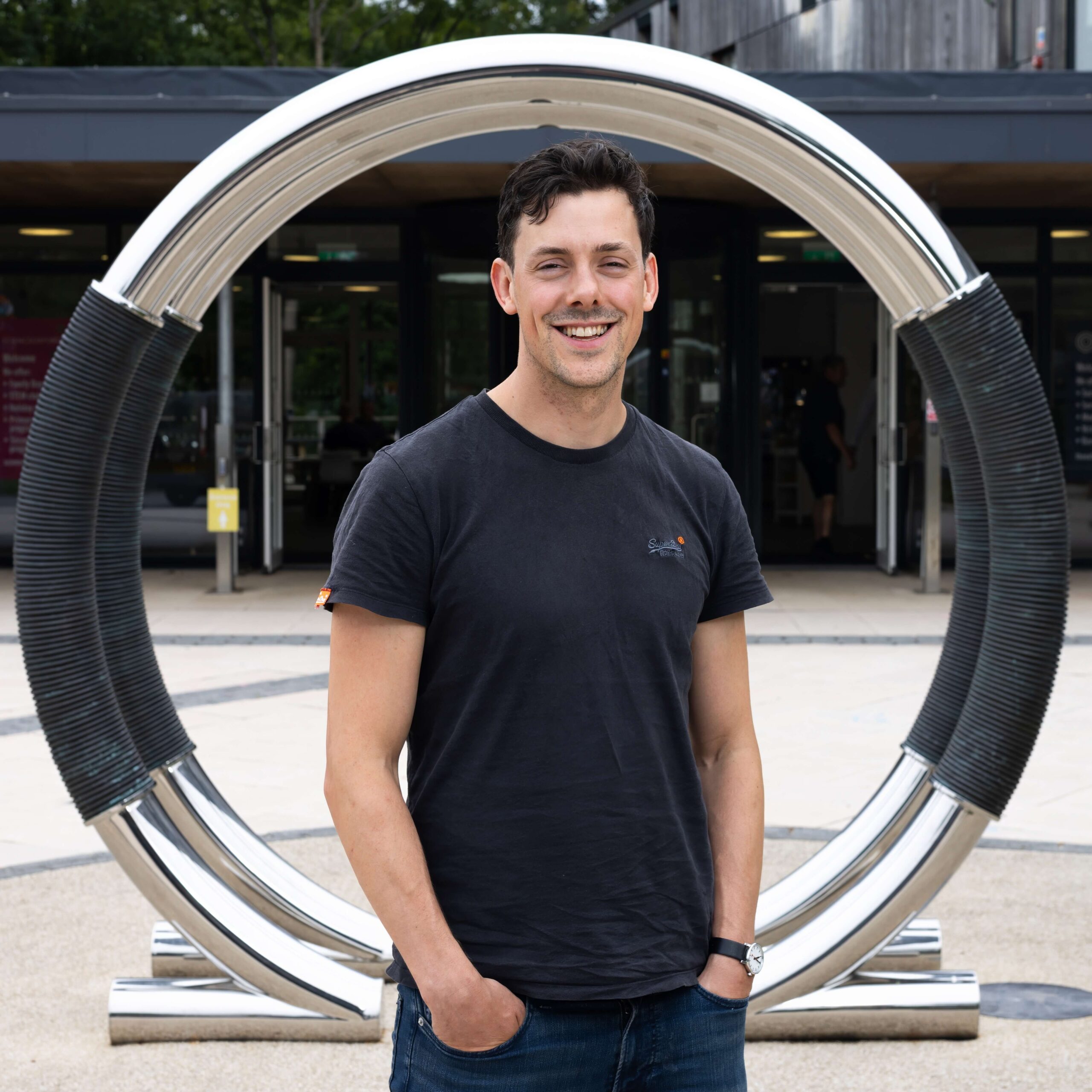 A man in a black t-shirt looks at the camera smiling. He stands behind a sculpture outside the Wood Centre for Innovation.